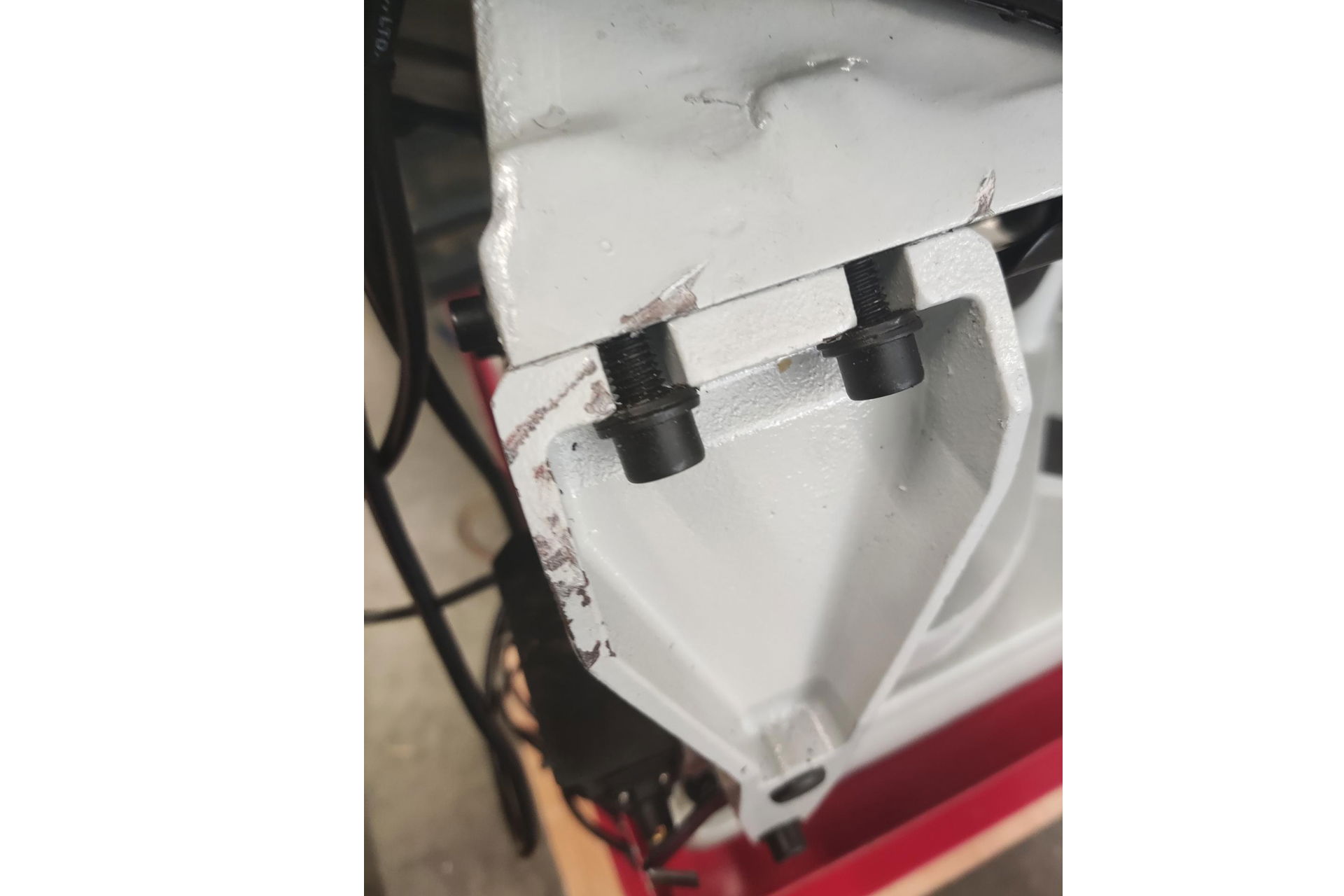 Detailansicht einer Werkzeugmaschine: Weiße, eckige Metallstruktur mit zwei schwarzen Schrauben. Kratzer in der Farbe sichtbar. Kabel im Hintergrund.